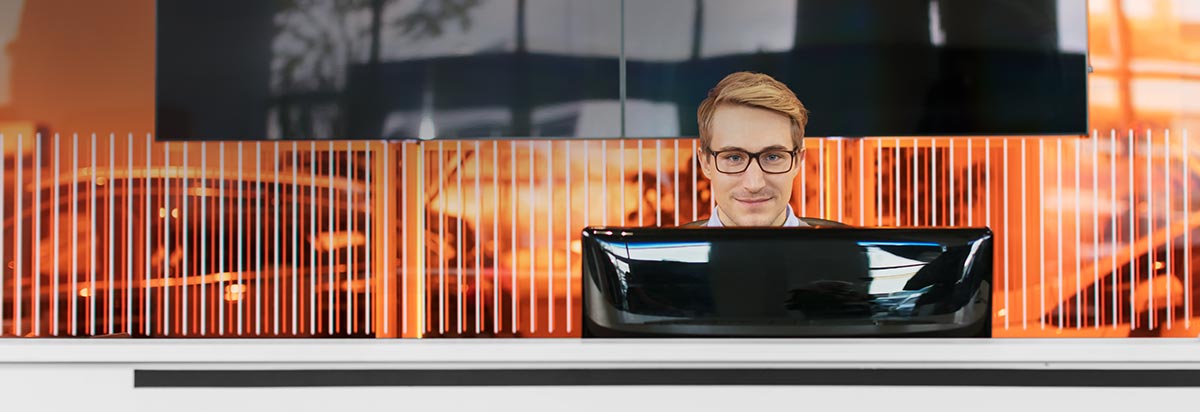 Ein junger Mann mit Brille sitzt hinter einem Empfangstresen, im Hintergrund warme Orangetöne.